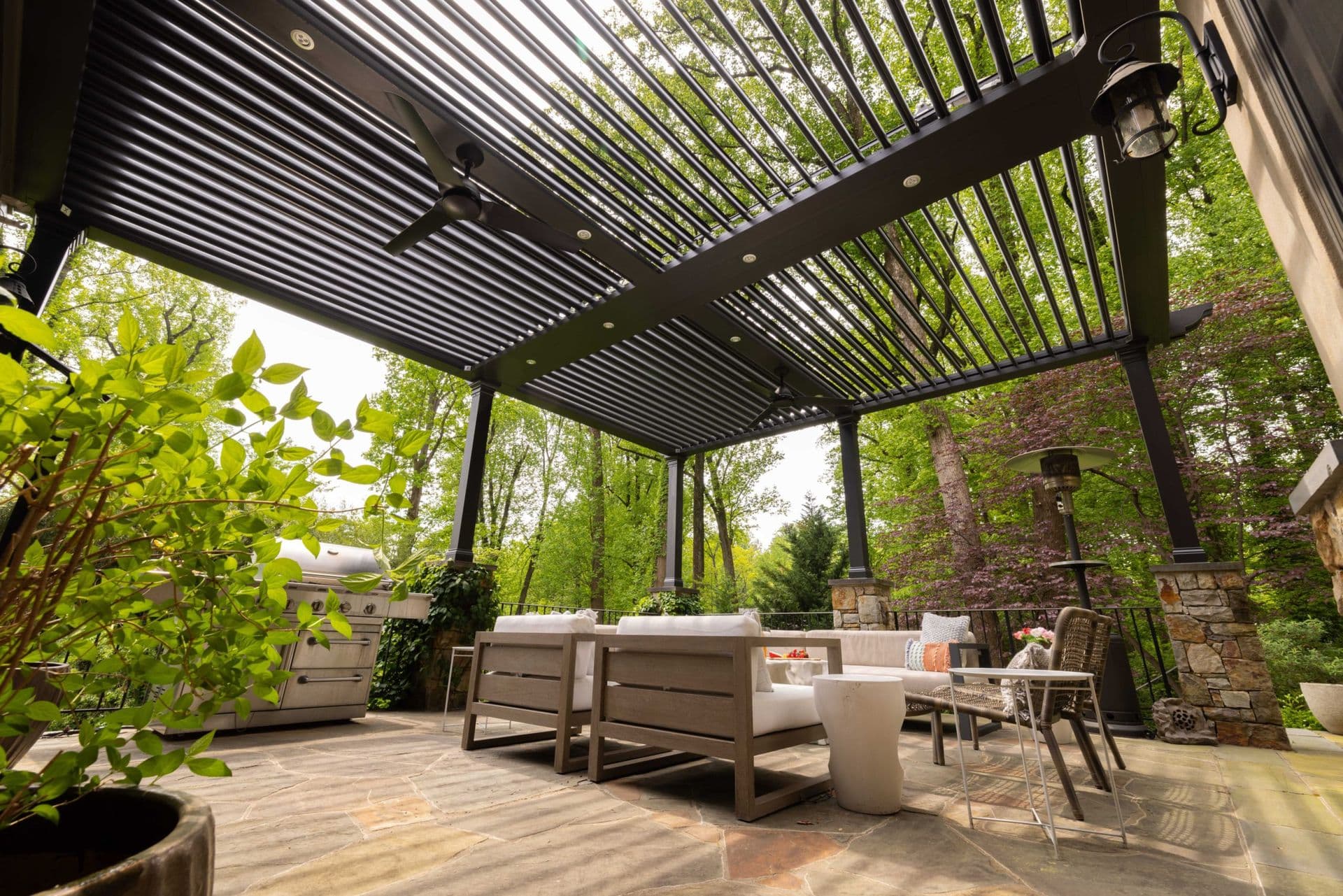 Covered outdoor patio with a modern slatted pergola, sofas, dining table, and lush trees, creating a serene, inviting retreat.