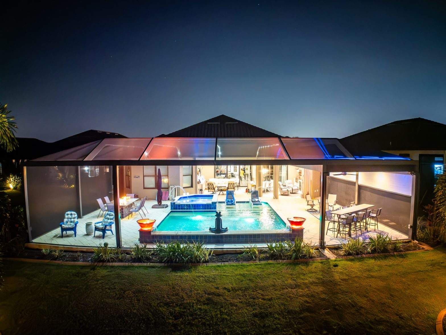 Nighttime view of a modern glass-walled home with a glowing indoor pool, lounge furniture, landscaped lawn, and dark sky.