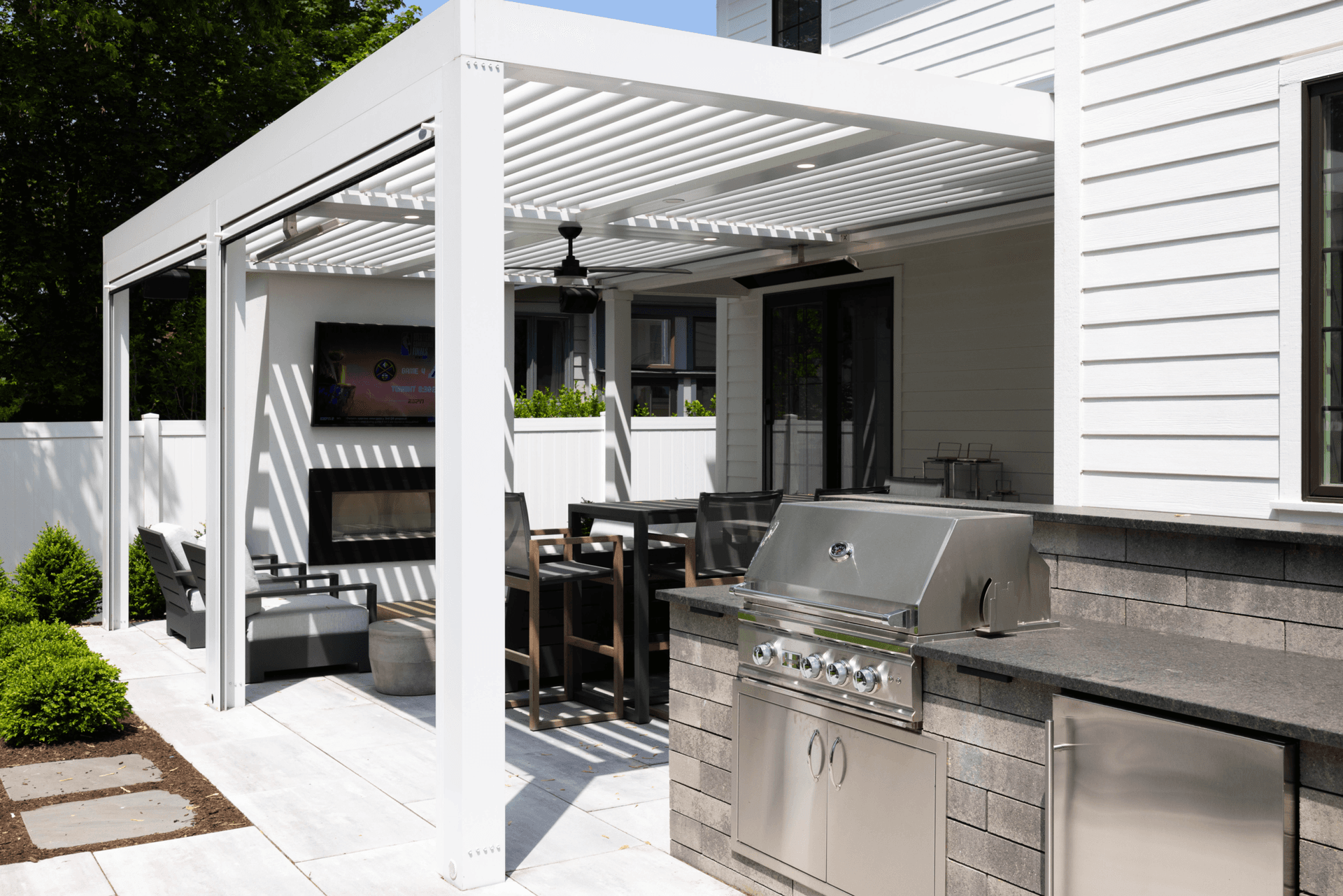 Modern white outdoor patio with pergola, ceiling fan, seating, stainless-steel grill, and stone counter in bright sunlight.