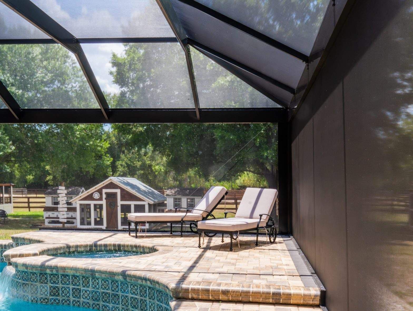 Sunlit screened pool enclosure with two lounge chairs, tiled deck, turquoise water, and a small backyard shed surrounded by trees.