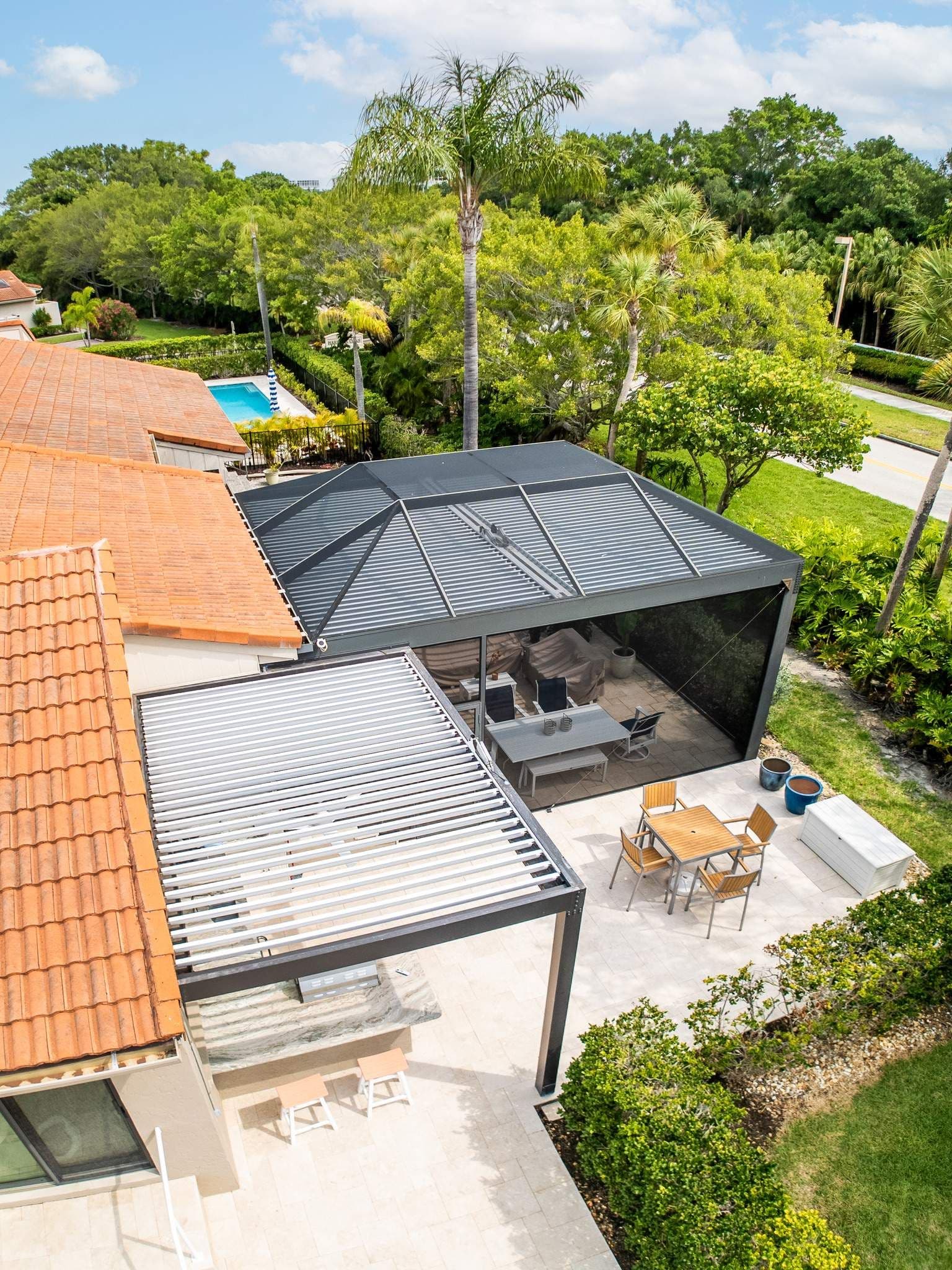 Aerial view of a tiled-roof home with covered patio, dining table, lounge seating, pool, lush tropical trees, and sunny blue sky.
