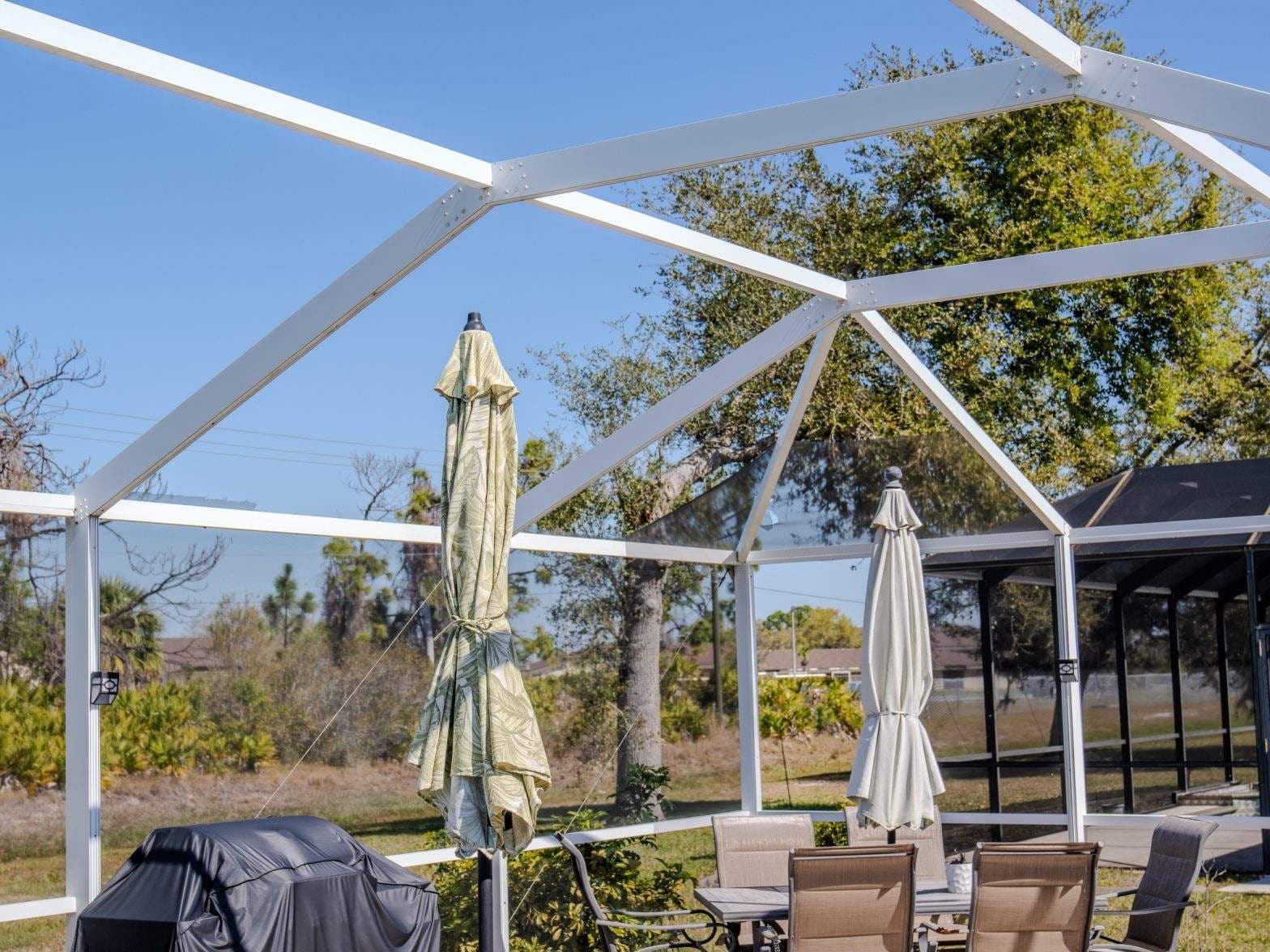 Outdoor patio with white pergola, closed umbrellas, covered grill, and seating overlooking trees and a sunny landscape under blue skies.