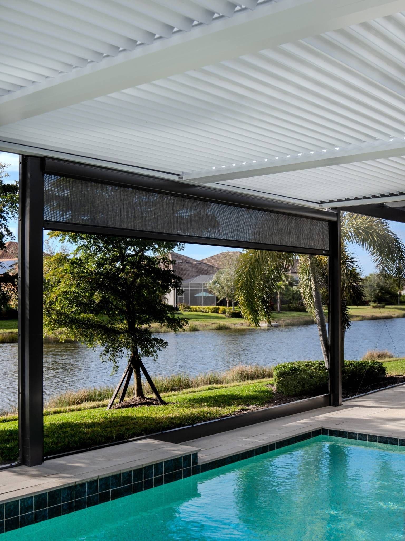 Covered patio with white pergola overlooks a turquoise pool, pond, landscaped lawn, trees, and homes in bright, tranquil sunlight.