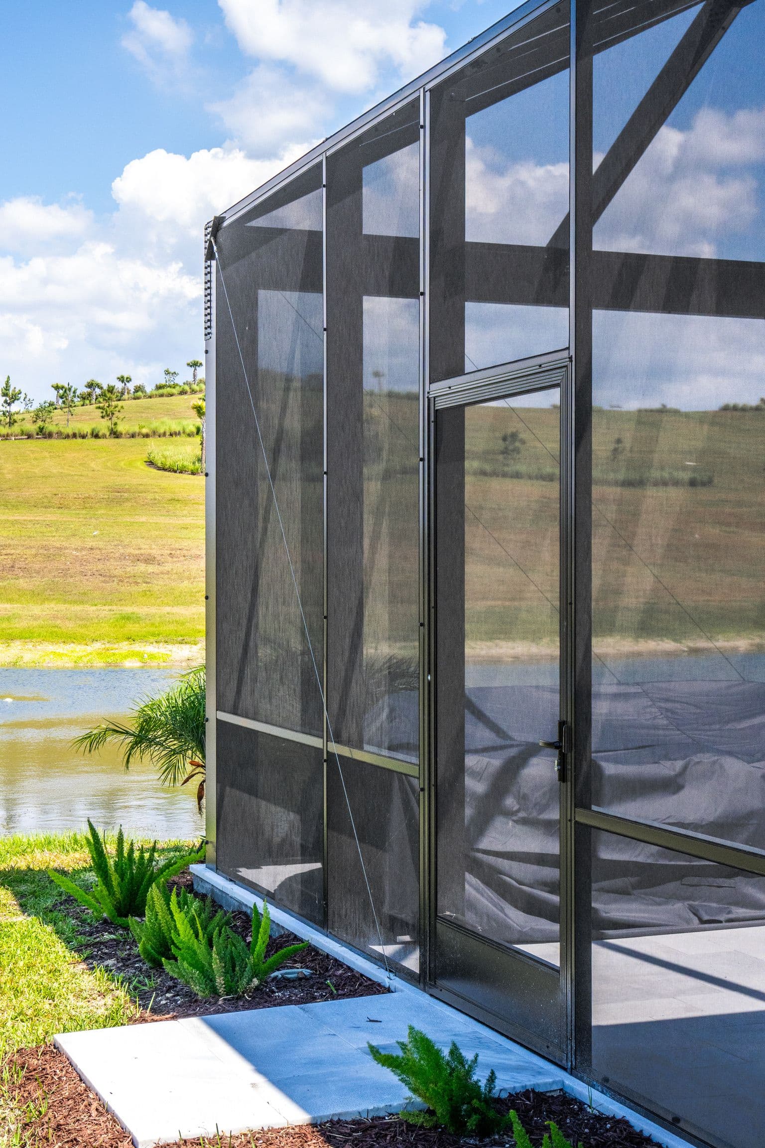 Modern black-framed screened patio enclosure beside a pond, with tropical plants, green fields, and blue sky in bright sunlight.
