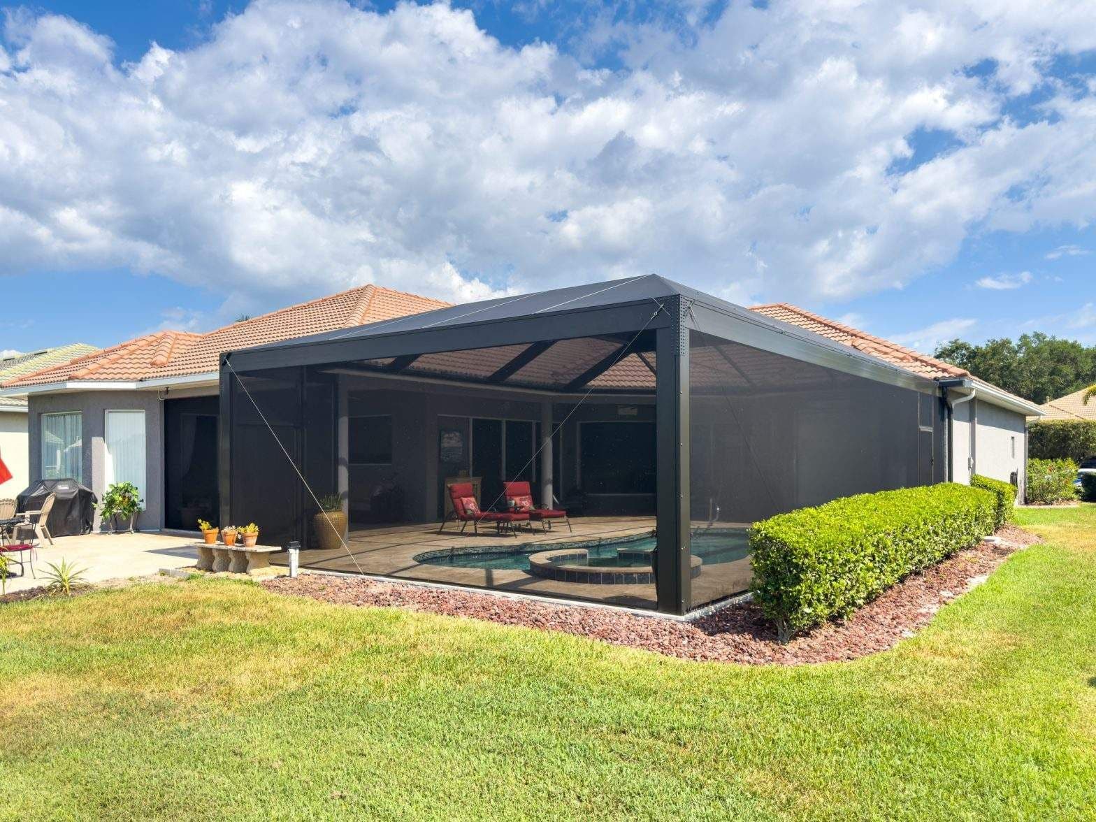 Sunny backyard with a screened-in patio, sparkling pool, red-tiled house, trimmed hedges, green lawn, and bright clouds creating an inviting mood.