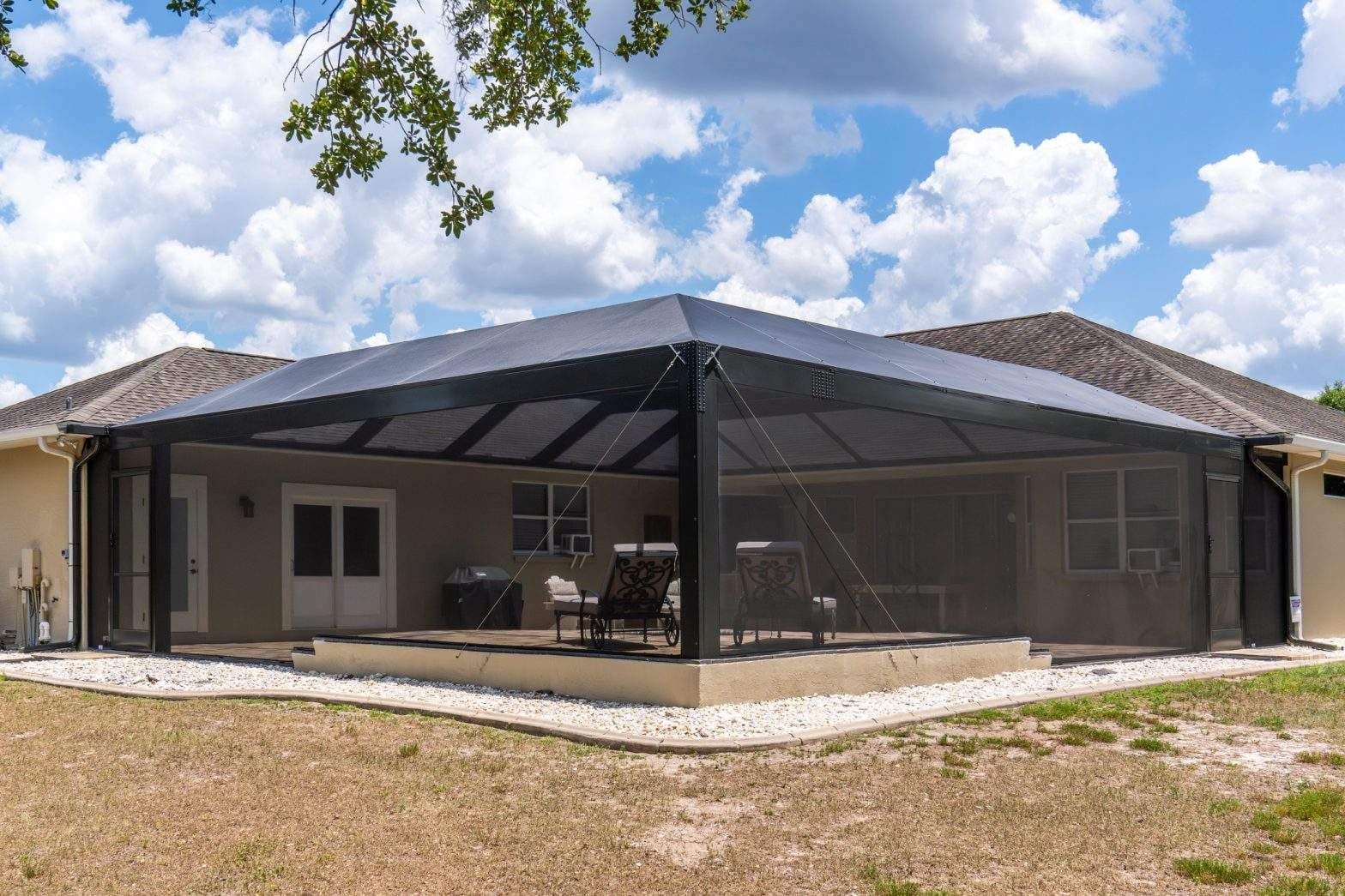 Single-story home with a large screened-in patio, outdoor seating, and covered terrace under a bright blue sky with fluffy clouds.