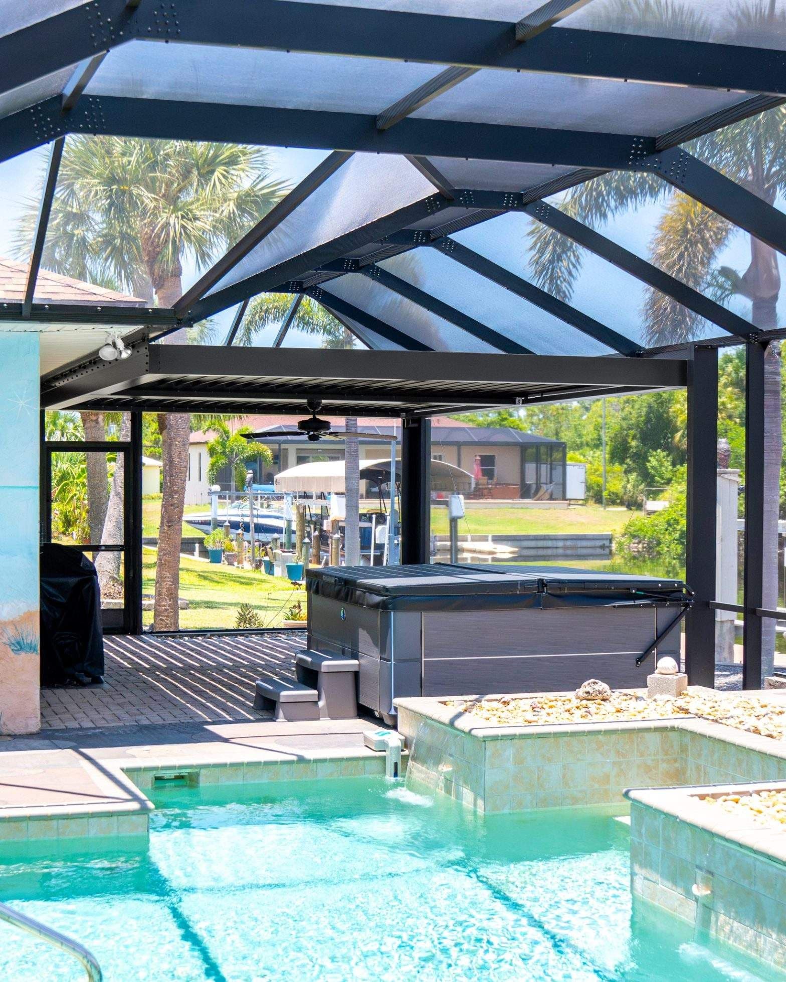 Sunlit outdoor pool beneath a black steel-and-glass canopy, with tiled fountains, palm trees, and a peaceful residential setting.
