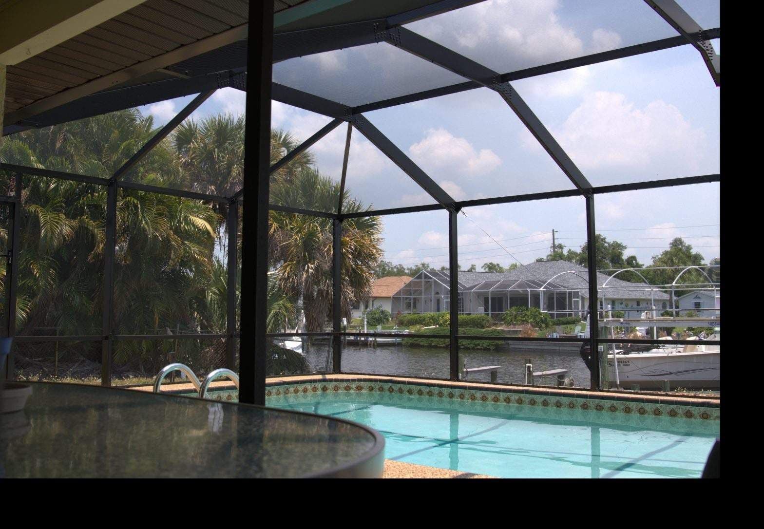 Screened-in backyard pool with turquoise water, patio table, and canal-side homes, framed by palm trees beneath a bright, partly cloudy sky.