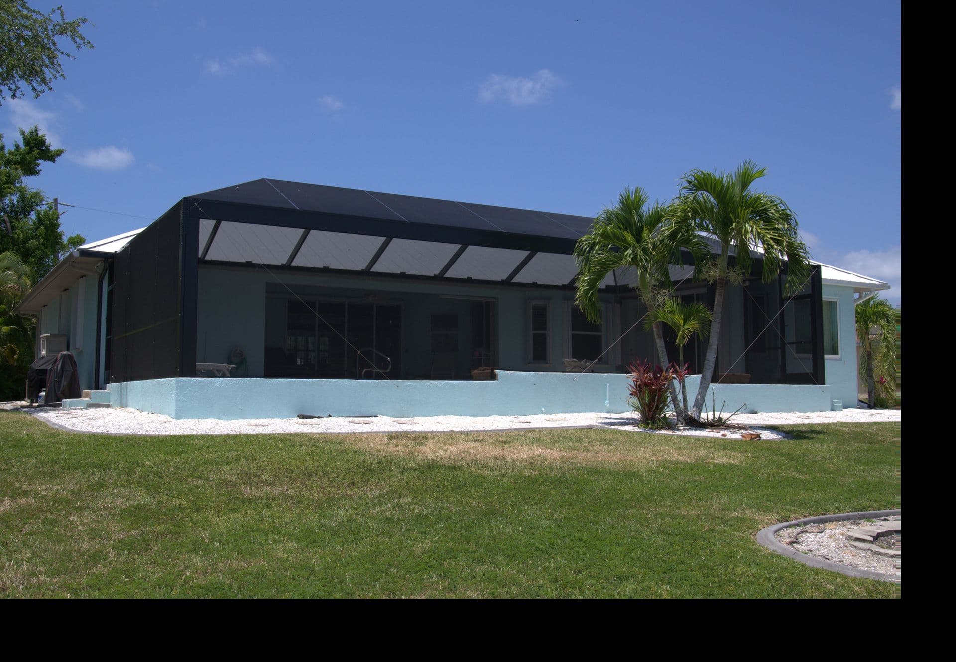 Modern single-story Florida home with a screened porch, palm trees, white gravel landscaping, and a green lawn beneath a bright blue sky.