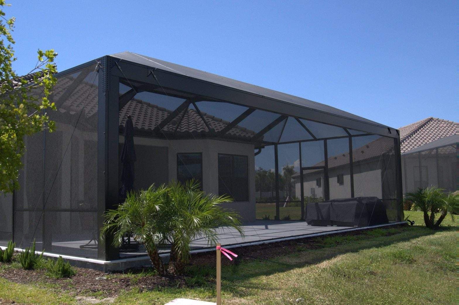 Screened patio enclosure beside a home, with dark framing, outdoor furniture, palm plants, and a sunny lawn creating a calm atmosphere.