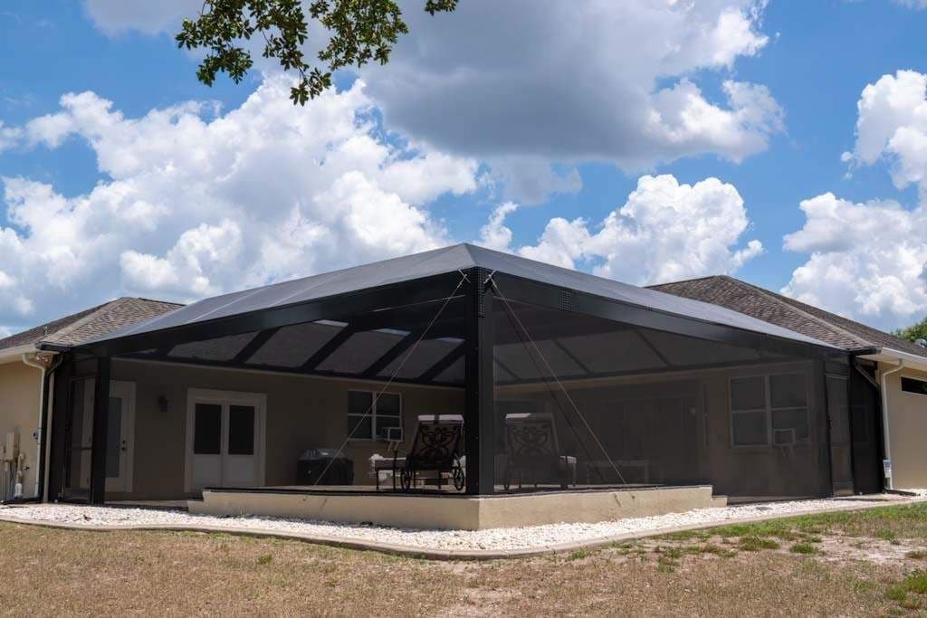 A spacious screened-in patio extends from a suburban home, furnished with chairs beneath a dark roof and bright clouds overhead, creating a calm setting.