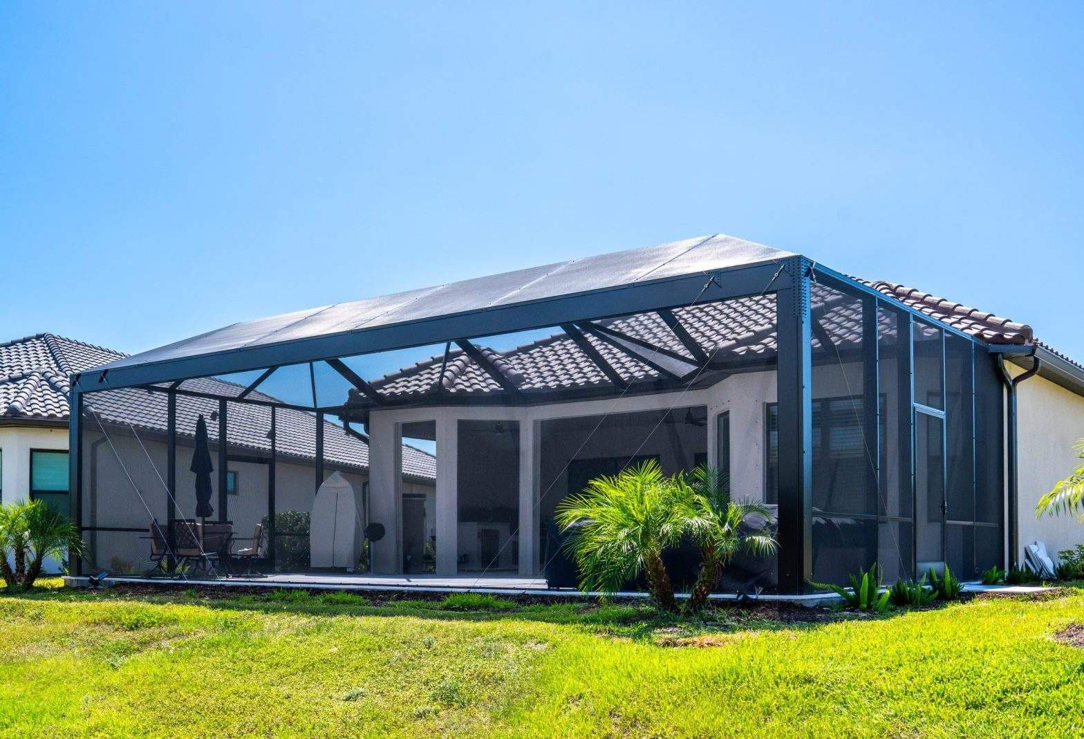 Sunny suburban home with a screened patio enclosure, glass doors, tiled roof, palm plants, and bright green lawn beneath a blue sky.