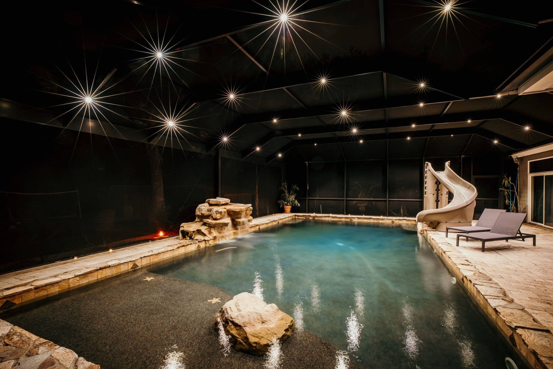 Luxury screened-in swimming pool at night with turquoise water, decorative starburst ceiling lights, stone accents, waterslide, lounge chairs, and fireplace.
