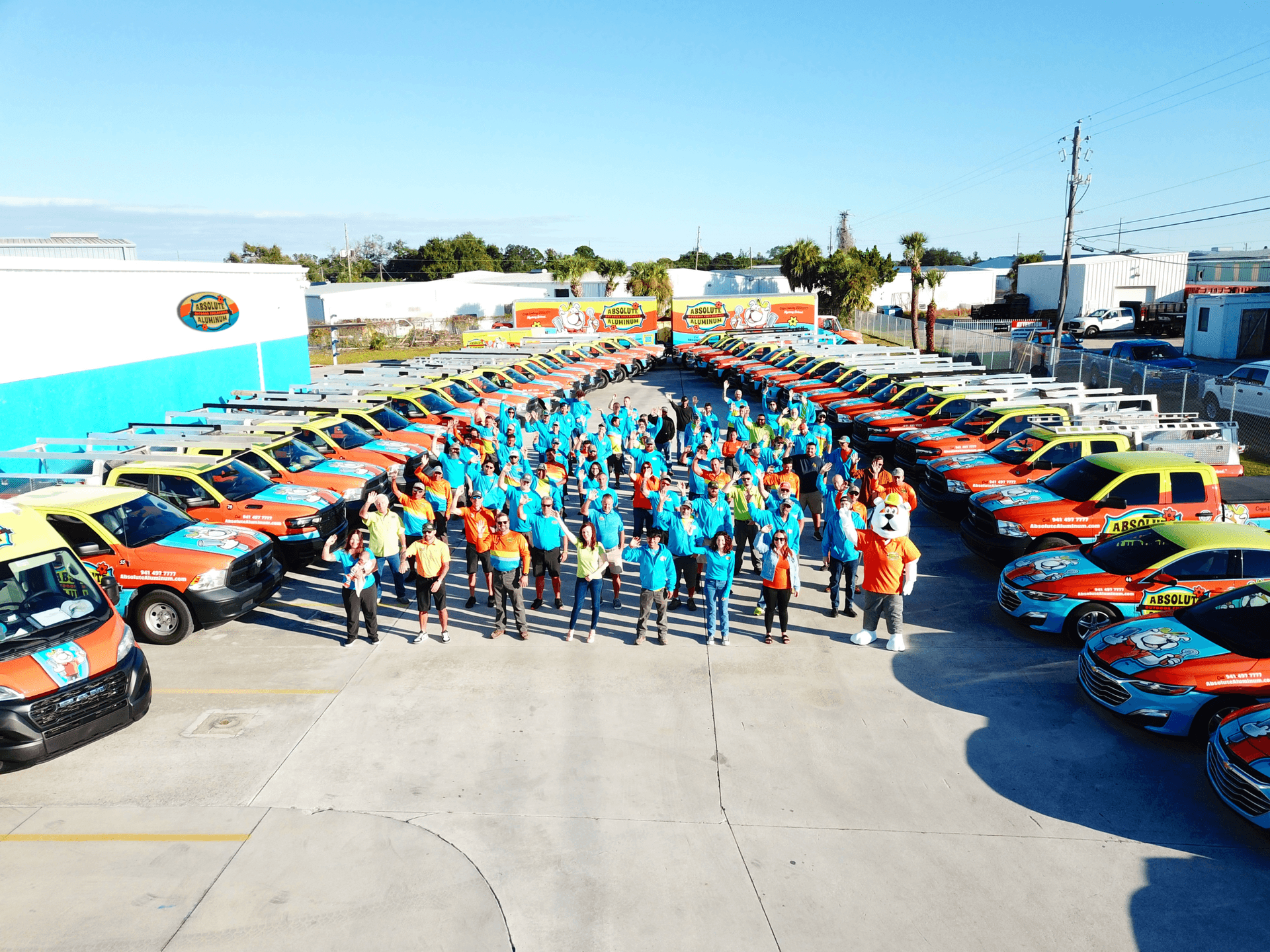 Large group of smiling employees in blue and orange shirts pose among rows of colorful service trucks in a sunny company parking lot.