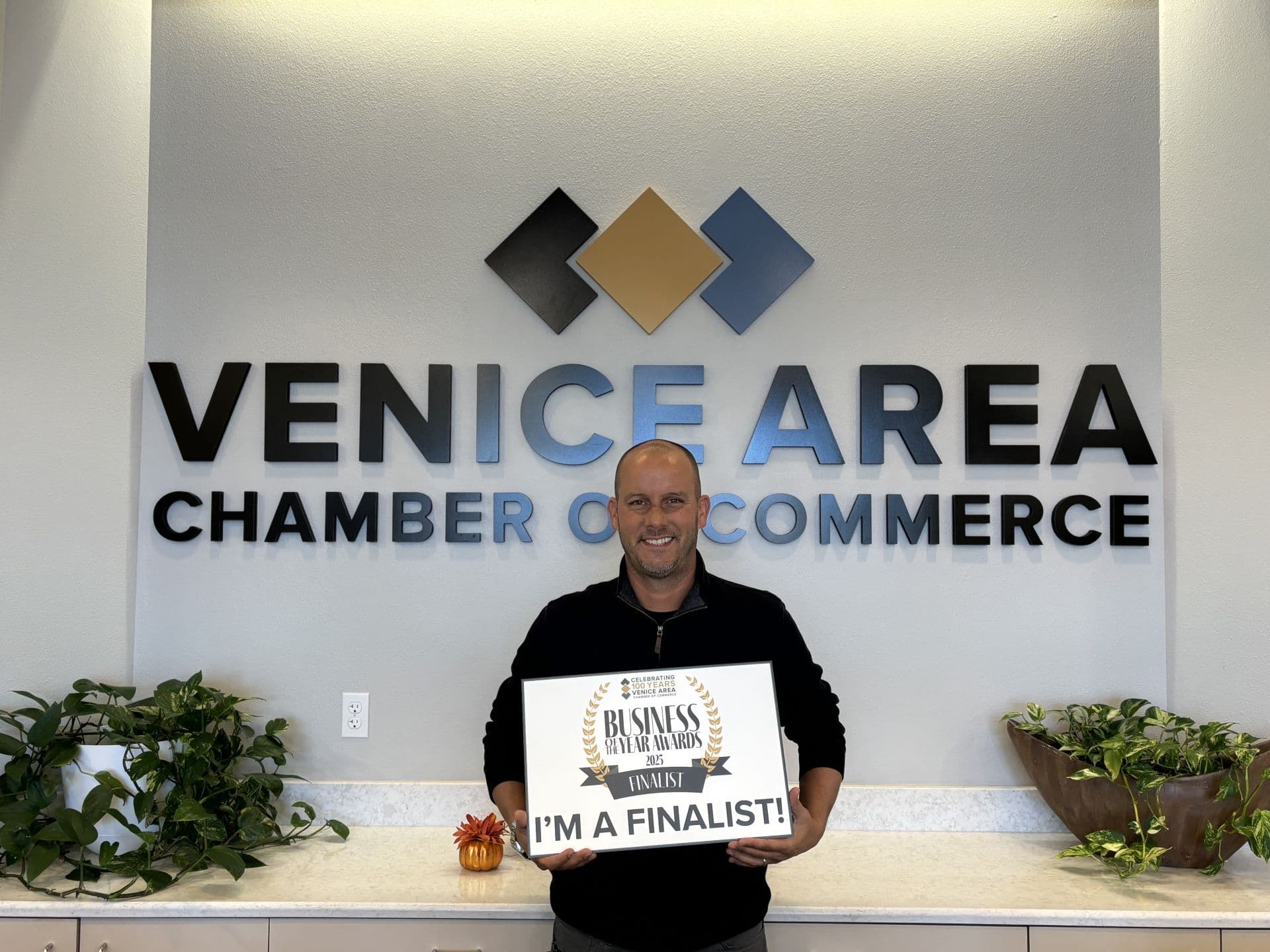 Smiling man holds an “I’m a finalist!” business award sign beneath the Venice Area Chamber of Commerce logo, conveying pride and celebration.