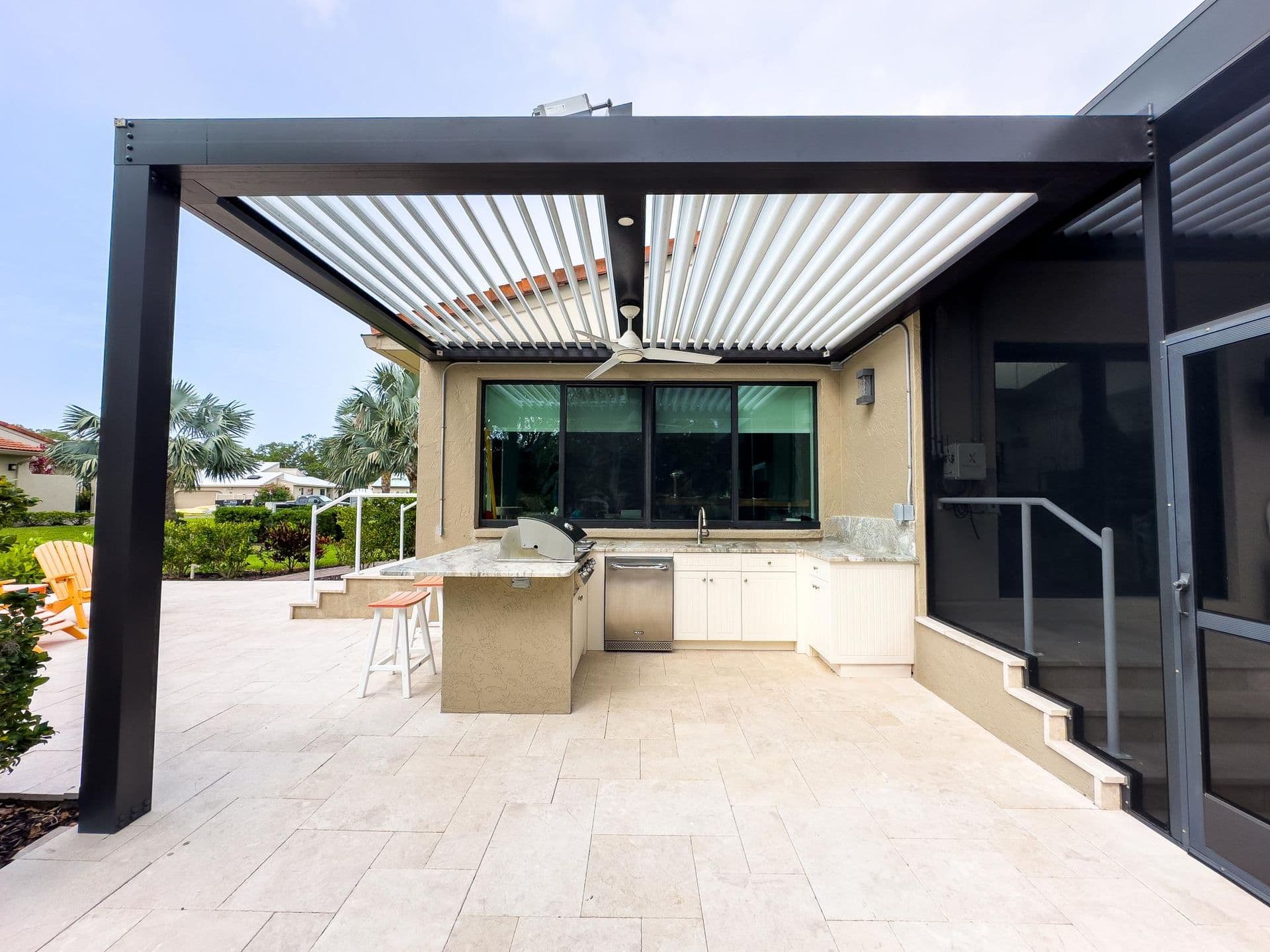 Modern patio with retractable louvered pergola over outdoor kitchen featuring grill, sink, and bar seating, adjacent to house