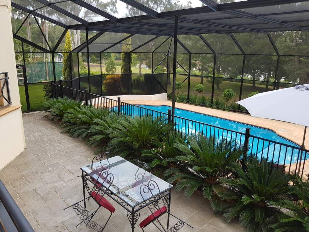 Screened backyard pool area with bright blue water, lush cycads, patio table and chairs, white umbrella, and peaceful greenery.