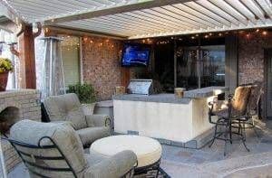 Covered outdoor patio with lounge seating, bar counter, and mounted TV, featuring brick walls and string lighting