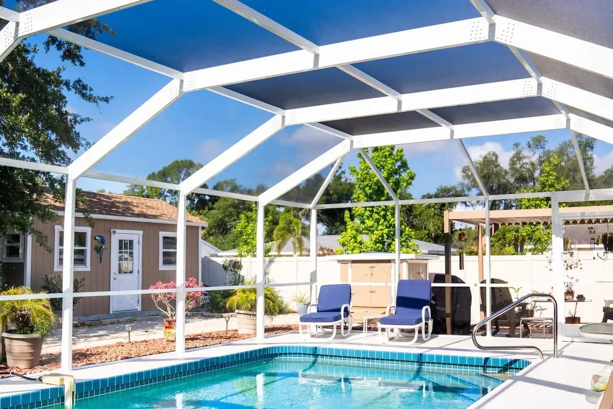 Bright screened-in backyard pool with blue water, lounge chairs, patio, tropical plants, and sunny skies creating a relaxing atmosphere.