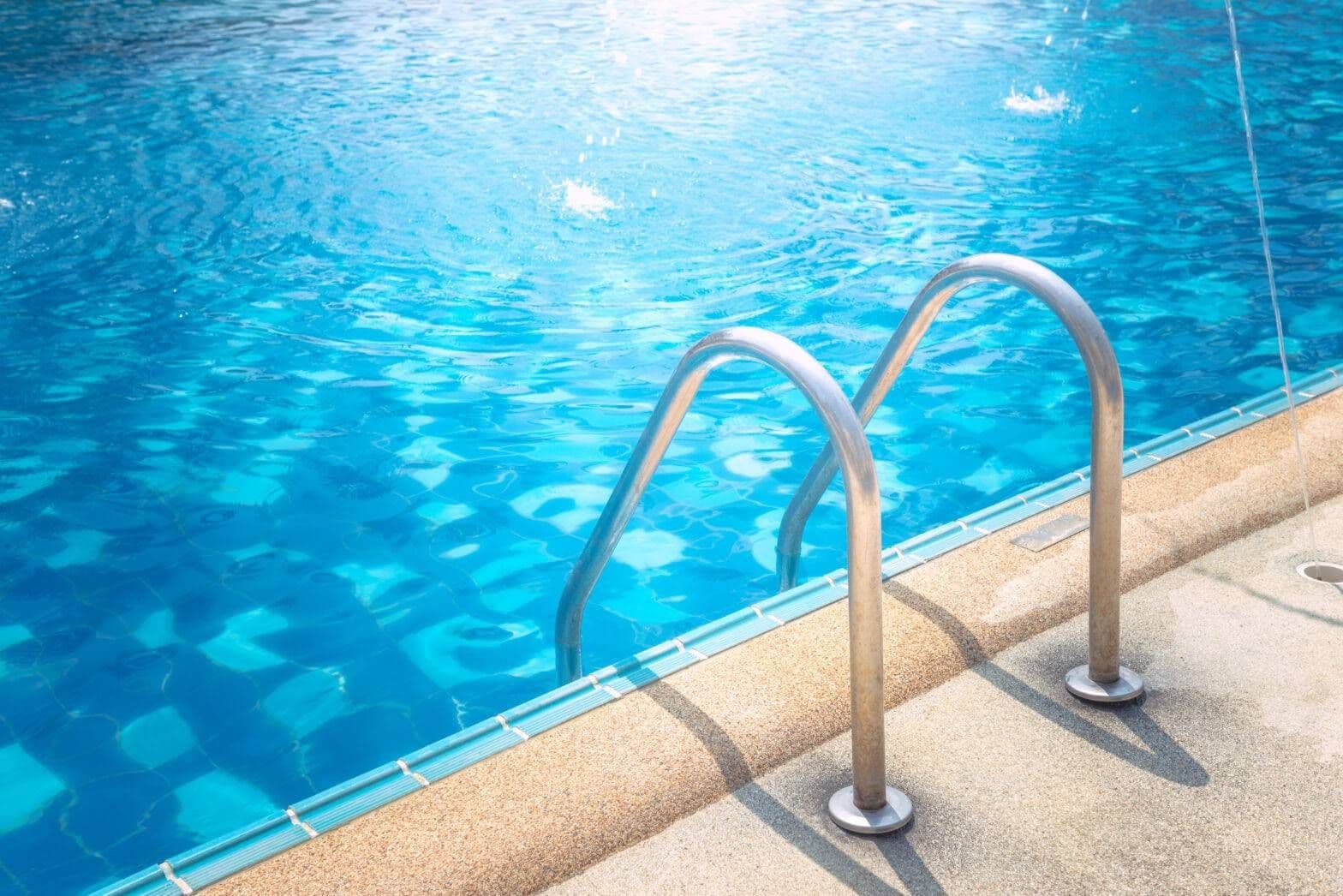 Bright blue swimming pool with a stainless-steel ladder, sunlight shimmering on the water and casting shadows across the deck.