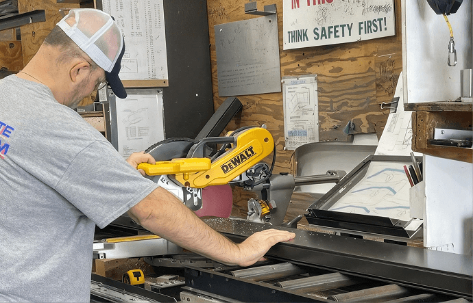 Man in a white cap using a DeWalt power saw in a workshop. Safety sign in the background. Focused atmosphere with tools and papers around.