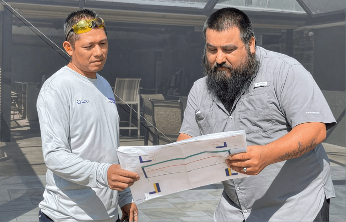 Two men examine construction plans outdoors. One, in a gray shirt, points at the paper. Both appear focused, engaged in a detailed discussion.