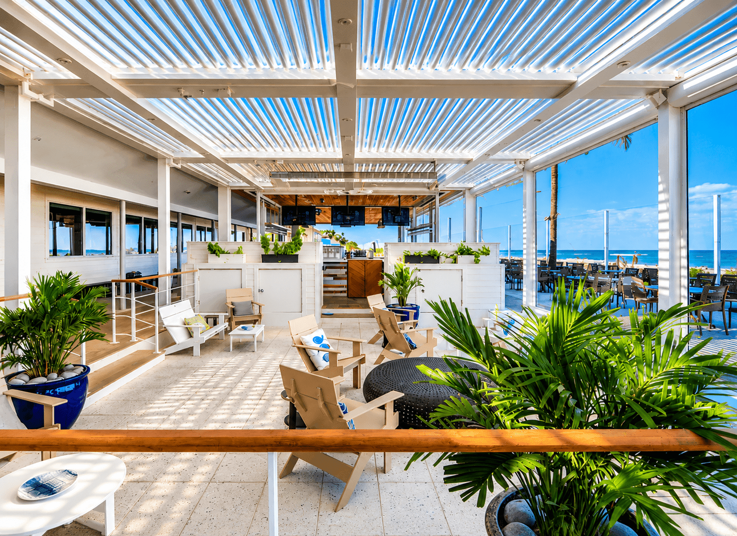 Modern beachfront restaurant and lounge with louvered pergola, wooden Adirondack chairs, potted tropical plants, and ocean views beyond.