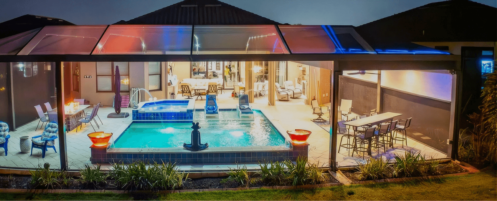 Modern home exterior at dusk with illuminated pool, spa, lounge areas, and bar seating visible through a screened pool and patio enclosure.