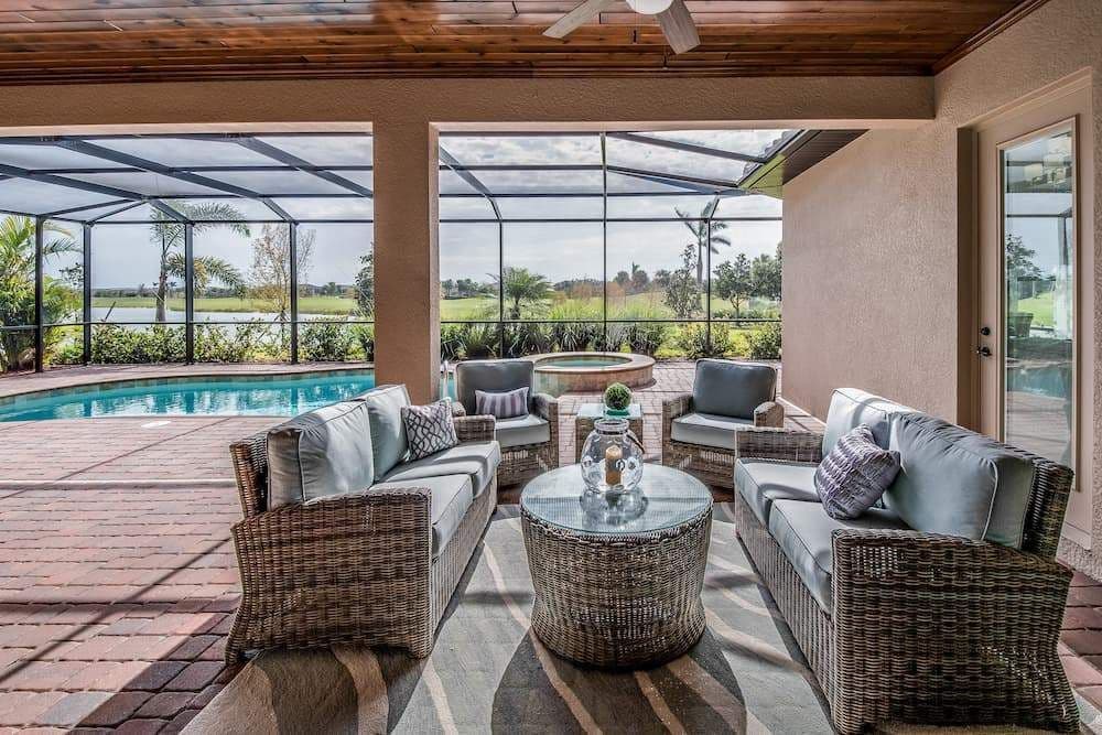 Covered outdoor patio with wicker sofas, chairs, and a round table overlooking a screened pool, palm trees, and sunny landscaped grounds.