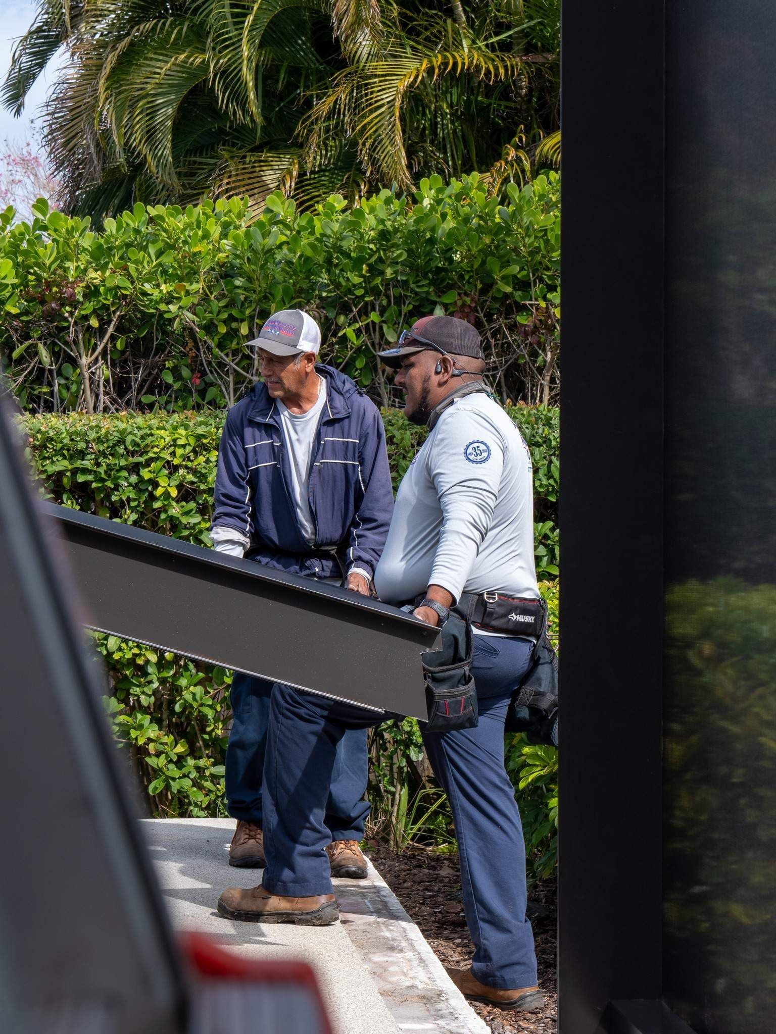 Two construction workers in safety gear carry a large steel beam past lush tropical hedges and palm trees on a sunny day.