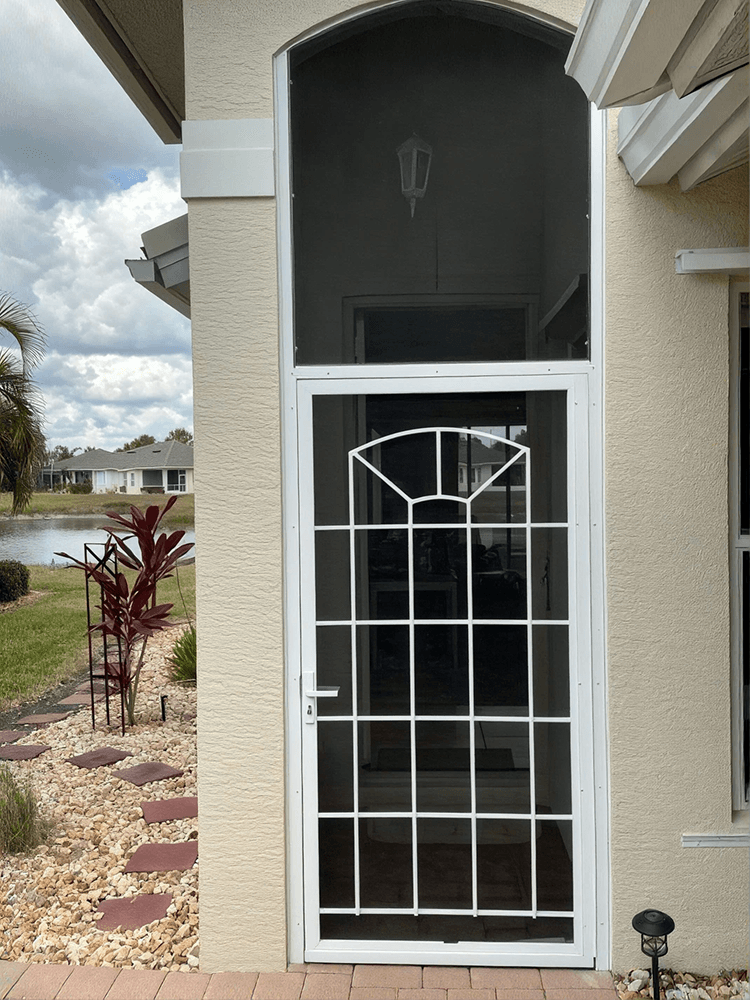 White stucco home entrance with a black security screen door, decorative white grille, stone landscaping, palm plant, and lake view.