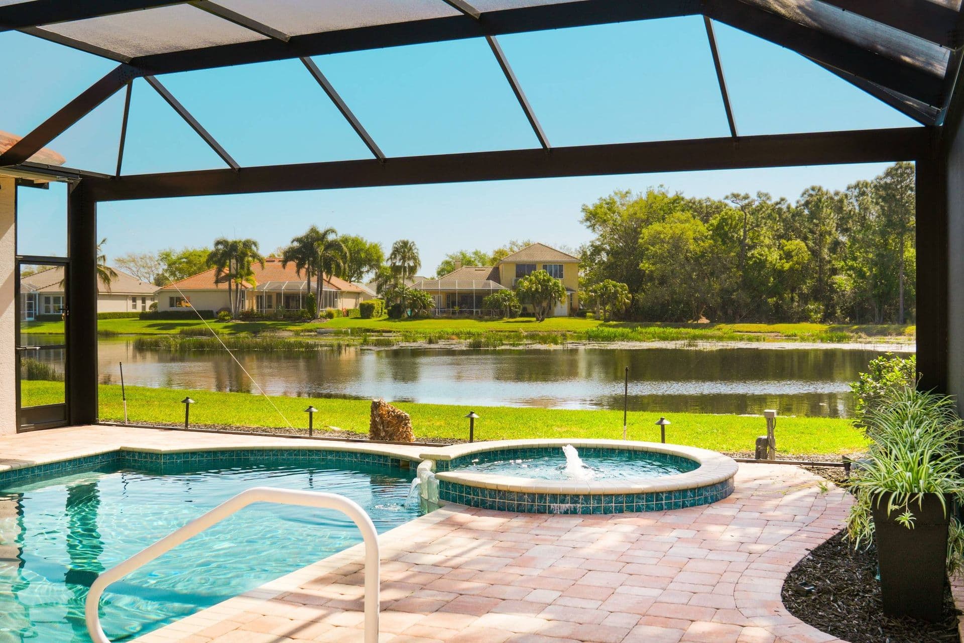 Covered patio with a pool and hot tub overlooks a tranquil pond. Bright blue sky, lush greenery, and distant houses create a serene, relaxing atmosphere.