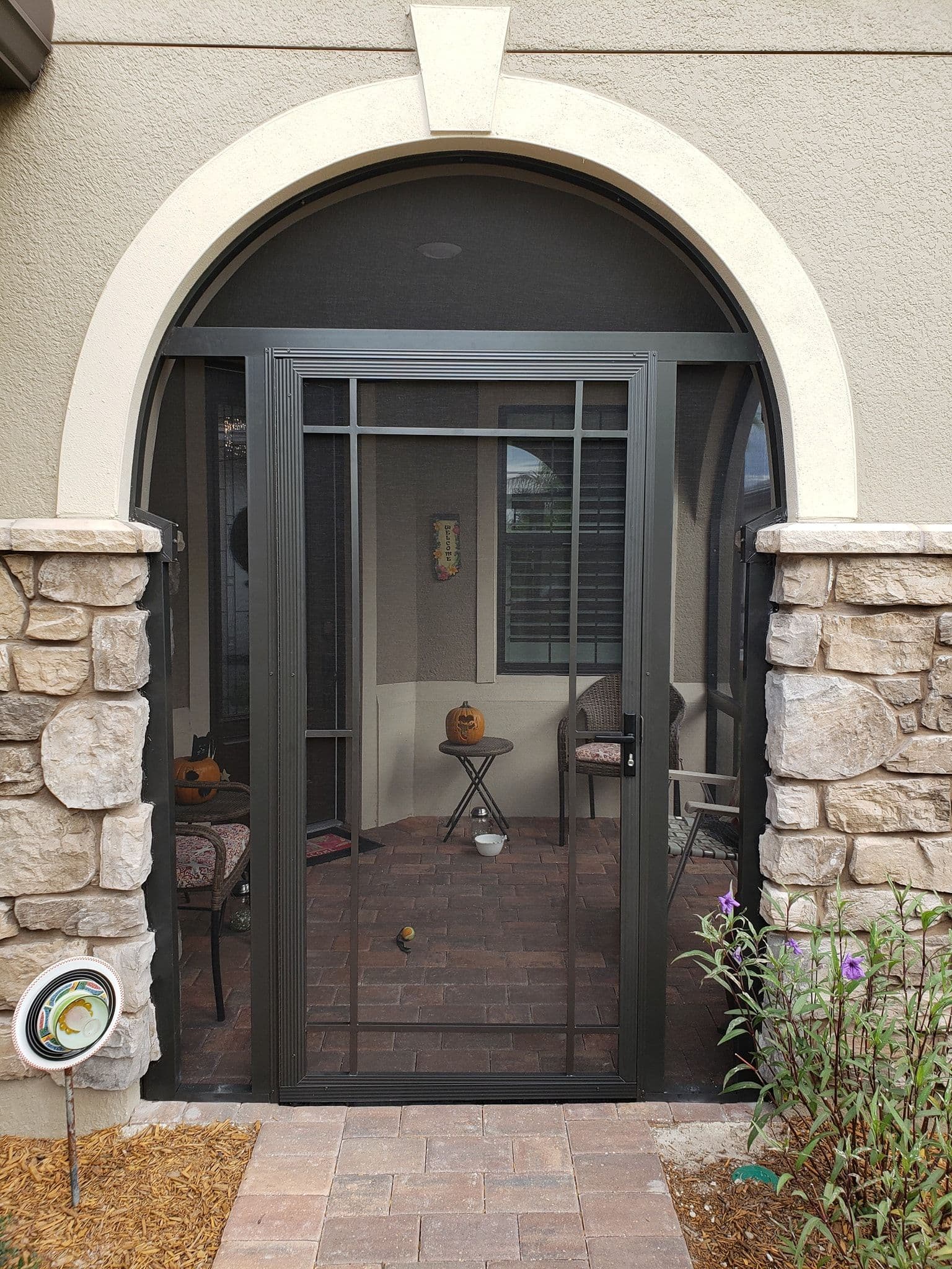 Arched stucco entryway with black screen door, stone pillars, brick patio, and flowering plants creates a welcoming home entrance.