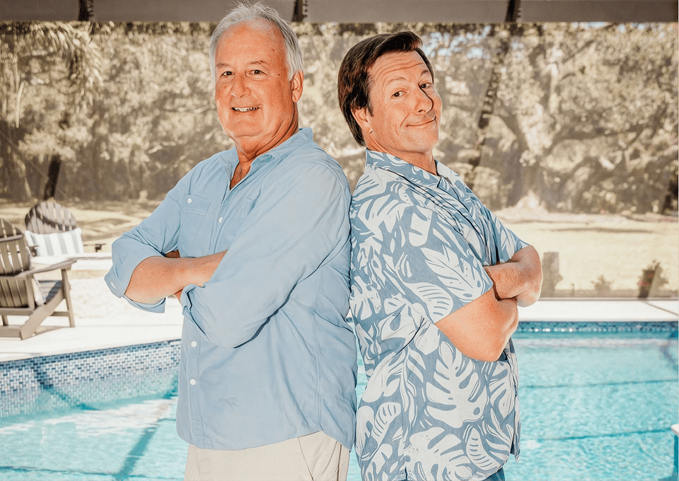 Two smiling men stand back-to-back with arms crossed beside a swimming pool, wearing casual shirts and projecting a playful, confident tone.