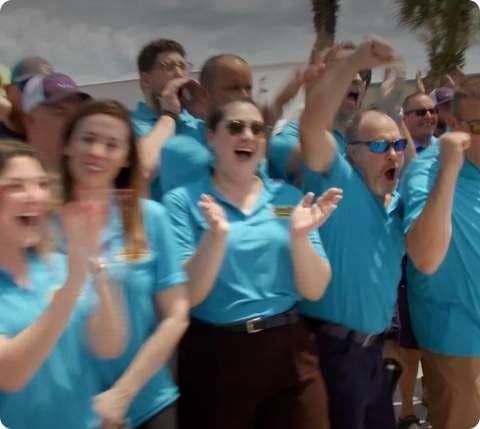 Group of people wearing blue Absolute Aluminum shirts cheering and celebrating outdoors with raised fists and joyful expressions.