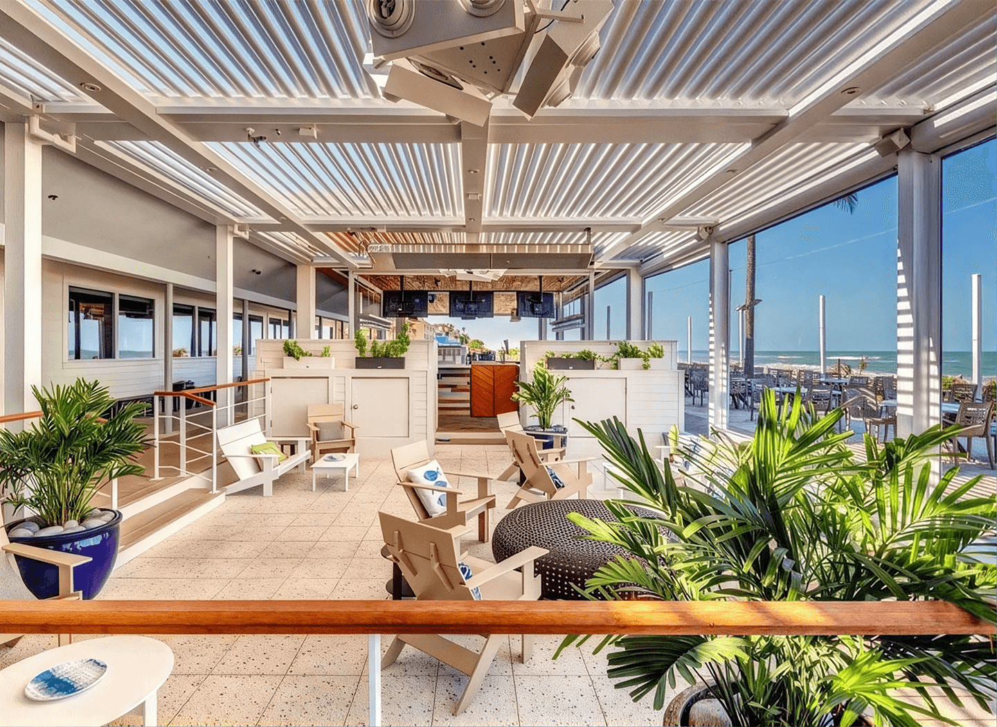 Bright oceanfront rooftop lounge with pergola, palm plants, wicker seating, and blue sea views, creating a sunny, relaxed atmosphere.