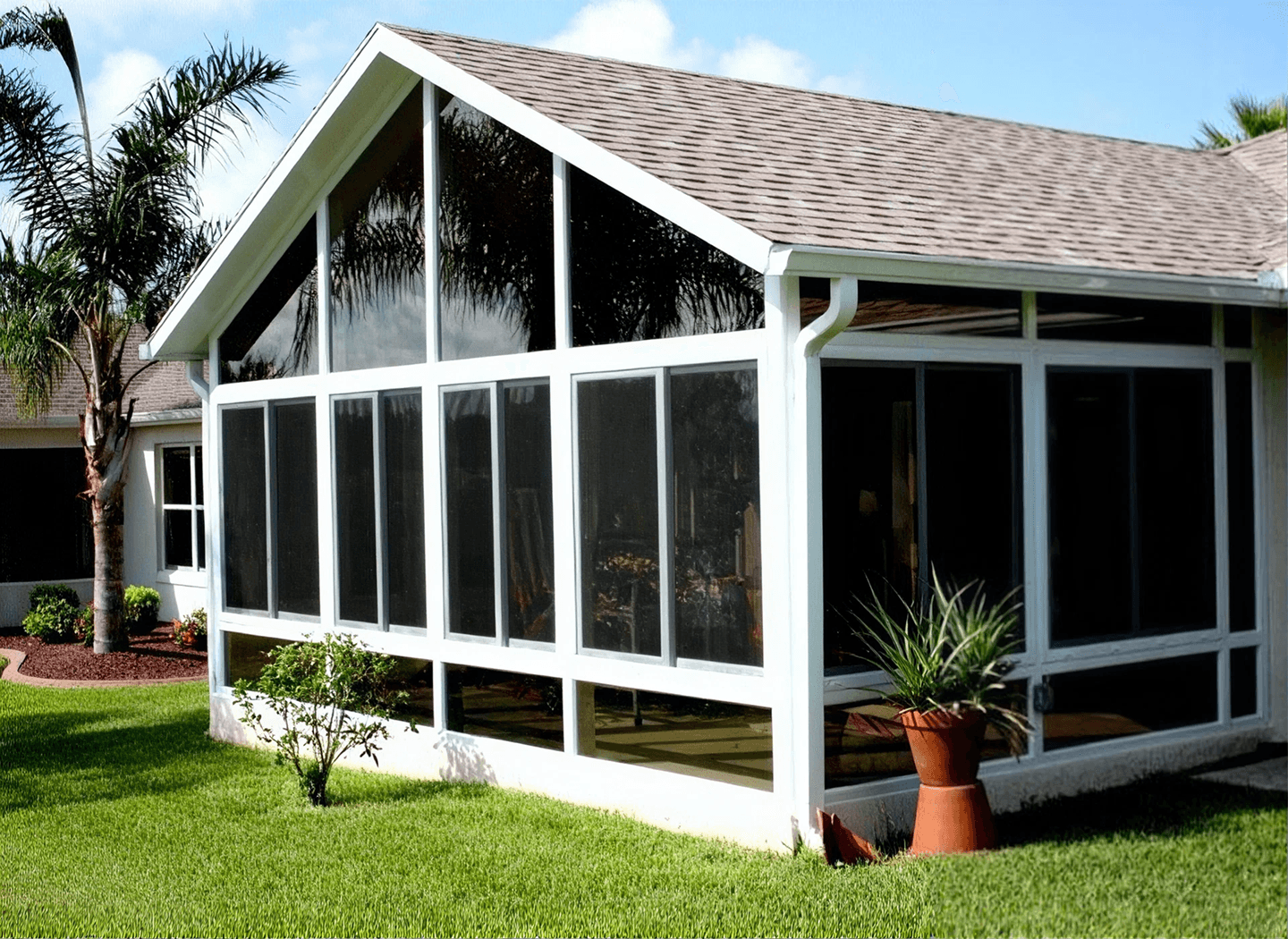 Bright Florida home with a screened-in patio, white framing, glass panels, palm trees, green lawn, and potted plants under a clear blue sky.