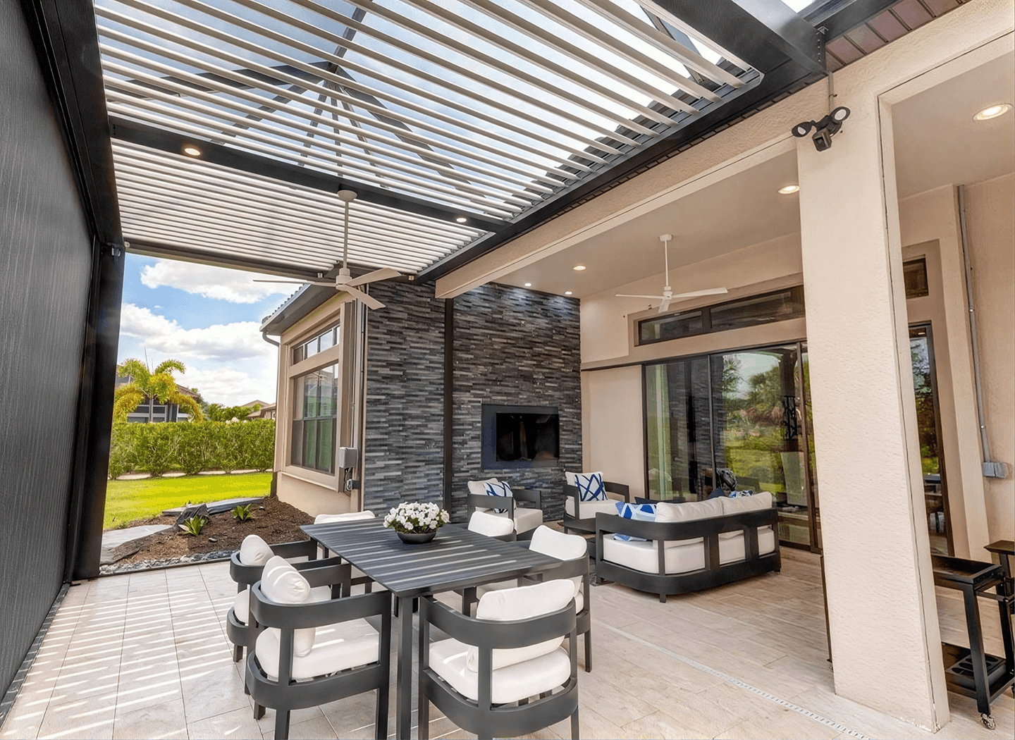 Modern outdoor patio with pergola roof, dining table, lounge seating, stone fireplace, glass doors, and lush garden views; bright and inviting.