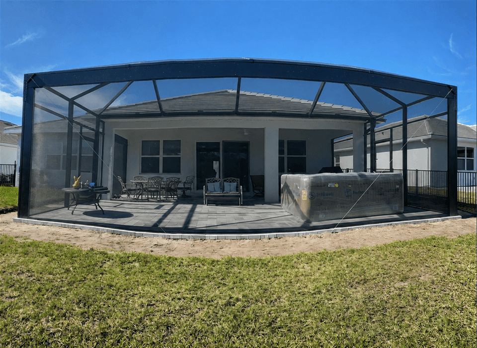 Screened-in patio enclosure with seating, grill, and pool equipment behind a suburban home, bordered by lawn under a bright blue sky.