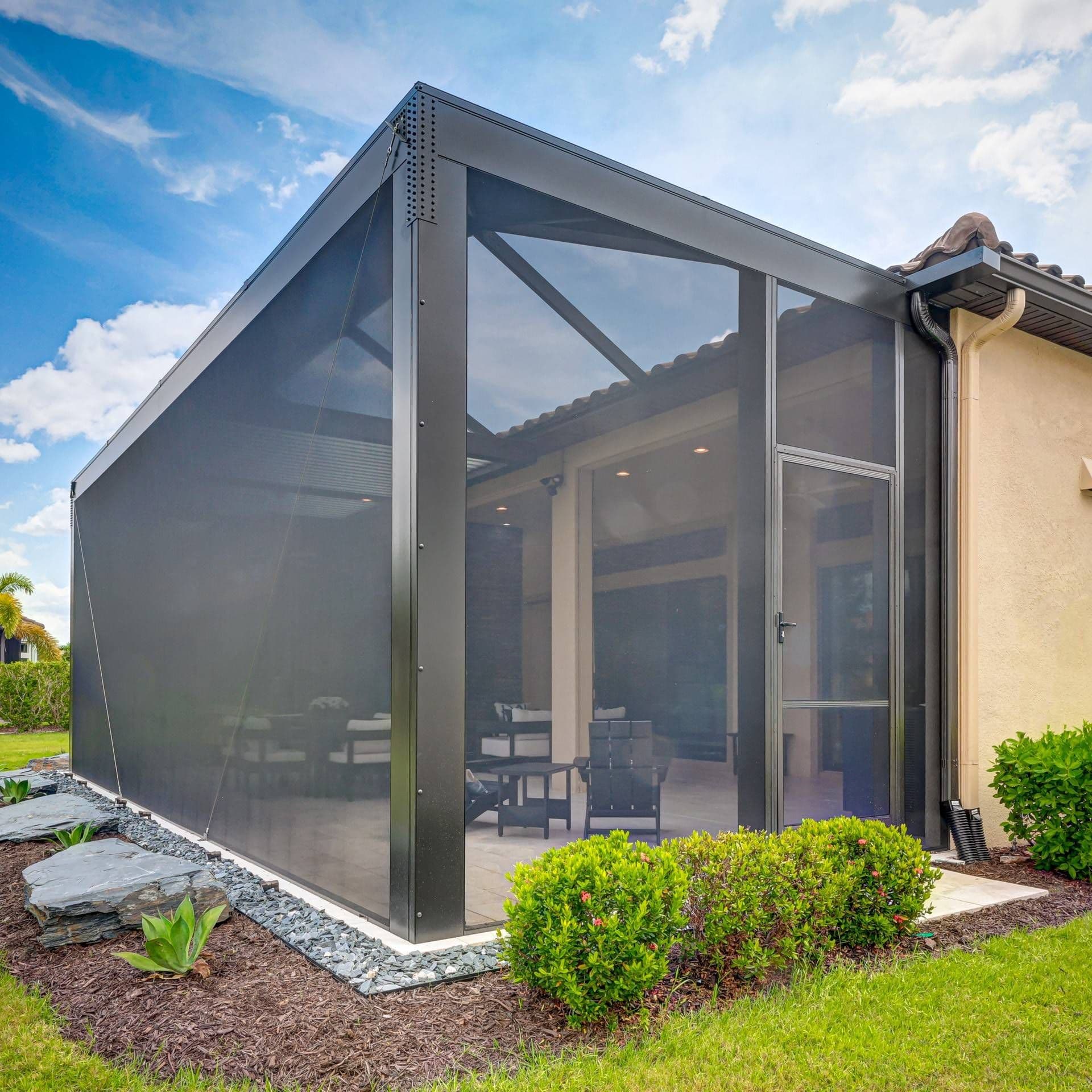 A screened patio with dark framing adjoins a beige house, surrounded by greenery. Inside, patio furniture is visible. The scene is sunlit and serene.
