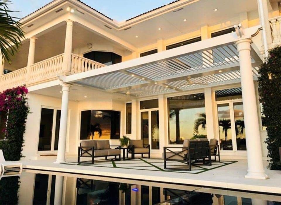 Elegant cream-colored villa with columns, balcony, covered patio, outdoor seating, and a reflective pool, creating a serene luxury setting.