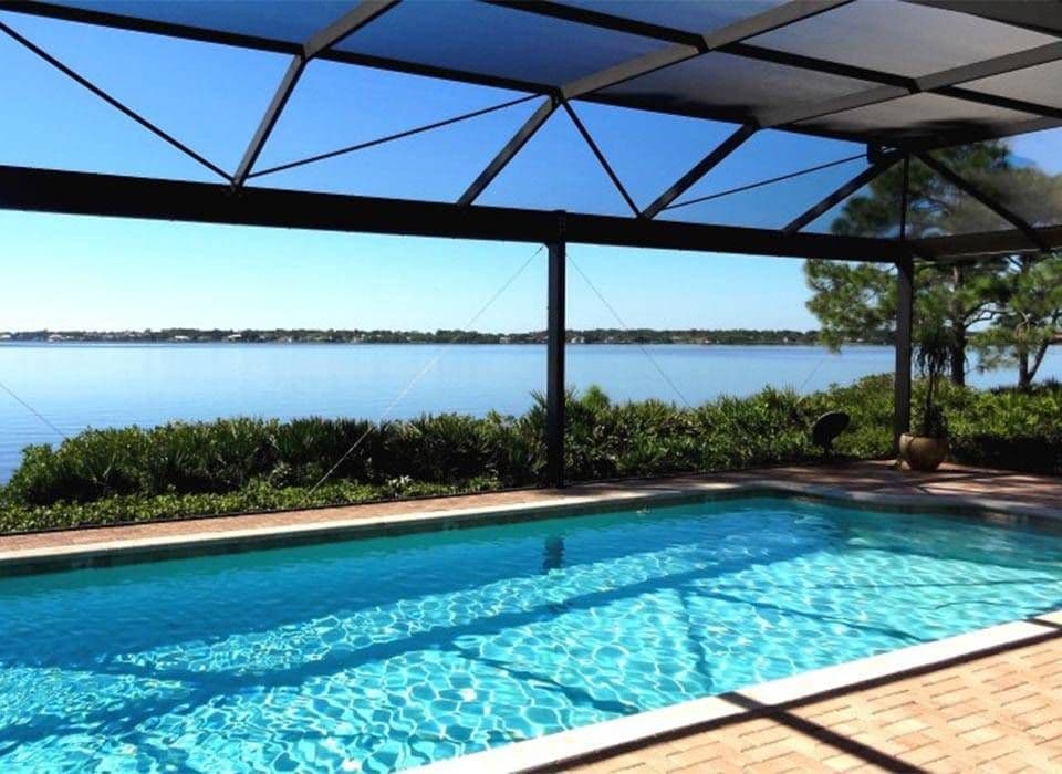 Sunlit backyard pool beneath a screened patio, with shimmering blue water, waterfront views, greenery, and a calm, relaxing atmosphere.
