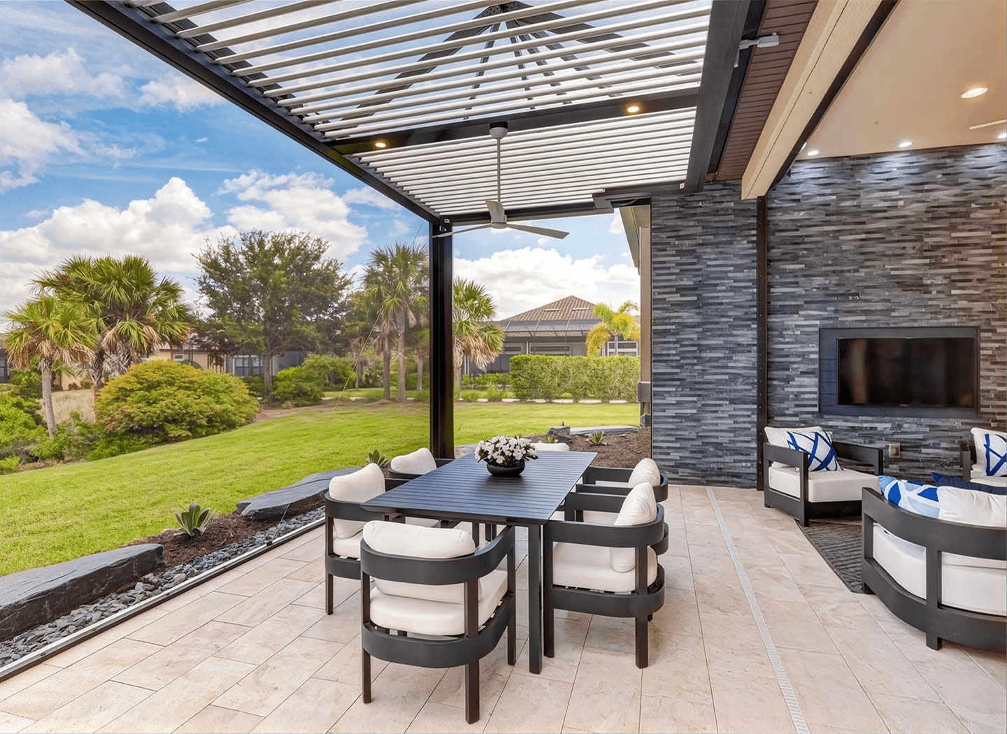 Modern outdoor patio with a sleek pergola, dining table, and cushioned chairs. Stone wall with a TV. Lush garden view and blue sky create a serene vibe.