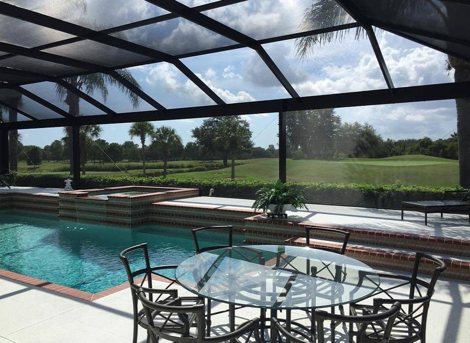 Sunlit screened patio with glass table and chairs overlooks a turquoise swimming pool, fountains, palms, and a green golf course.