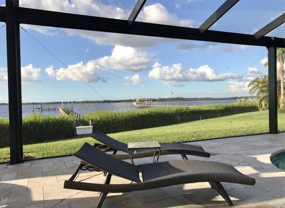 Poolside patio with black lounge chairs overlooks a calm lake, green lawn, dock, palm tree, and bright blue sky with clouds.