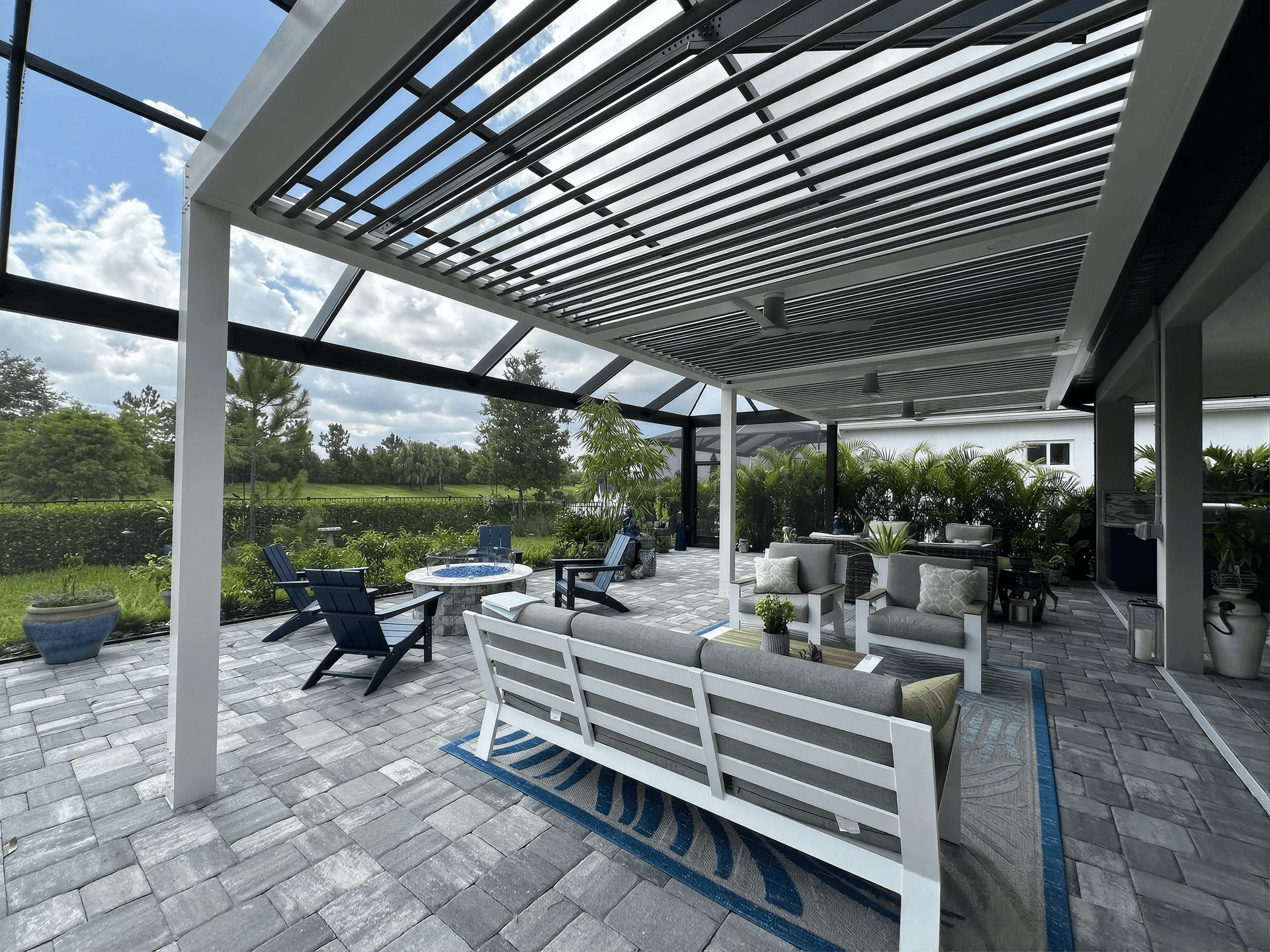 Covered patio with modern seating, a fire pit, and potted plants. Overhead slatted pergola provides shade. Greenery and open sky create a serene atmosphere.