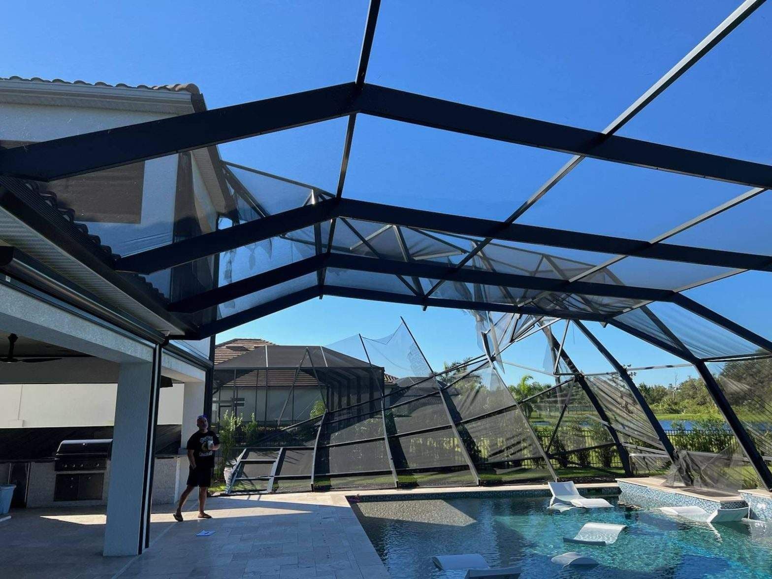 Backyard swimming pool beneath a damaged screened enclosure, with bent black beams and torn mesh; a person stands beside the house.