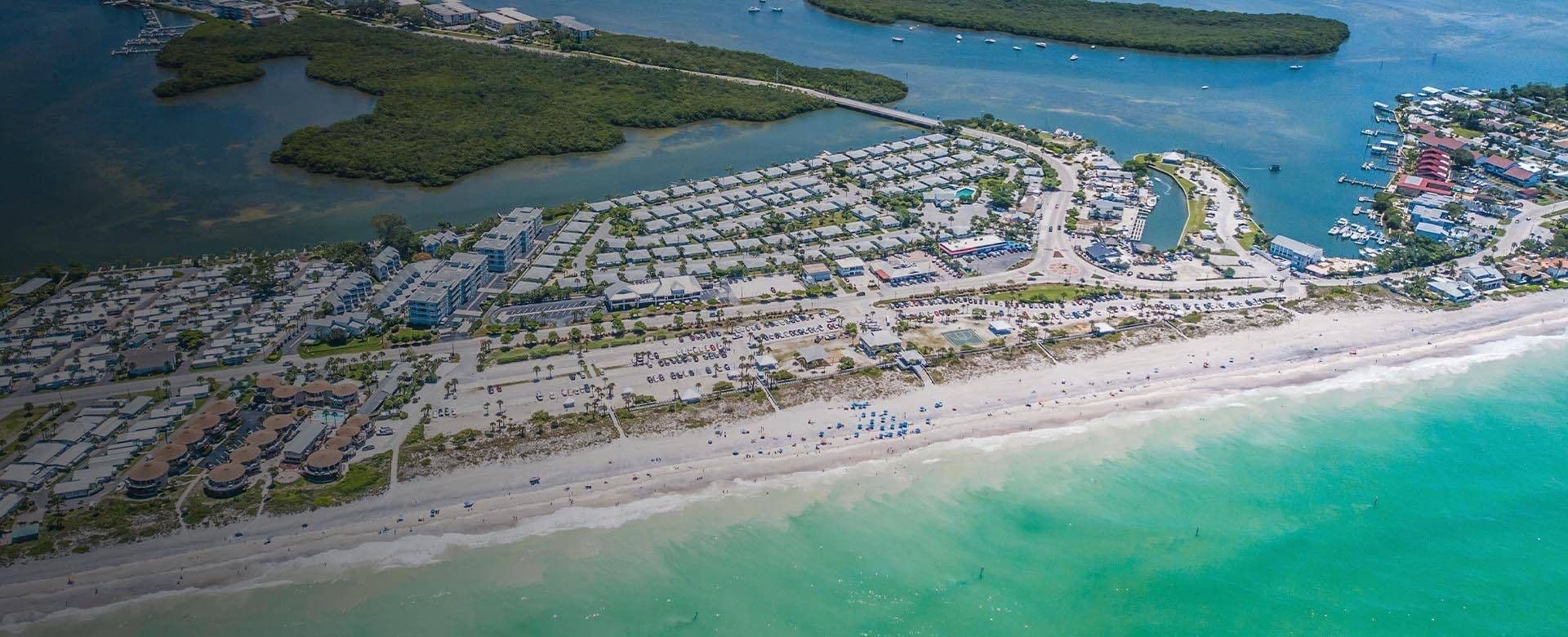 Aerial view of a beach with turquoise water, white sand, and a resort area featuring palm trees and sun loungers.