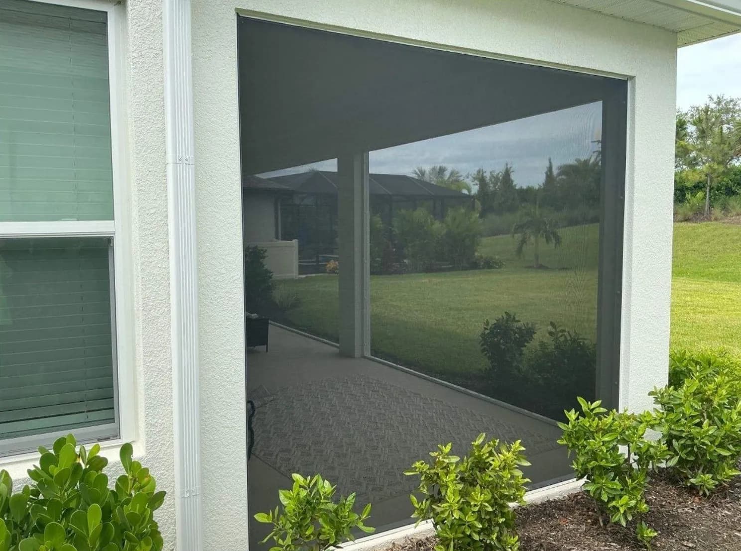 Screened-in patio with dark mesh panels, reflecting a green lawn and trees, bordered by white walls and neatly trimmed shrubs.