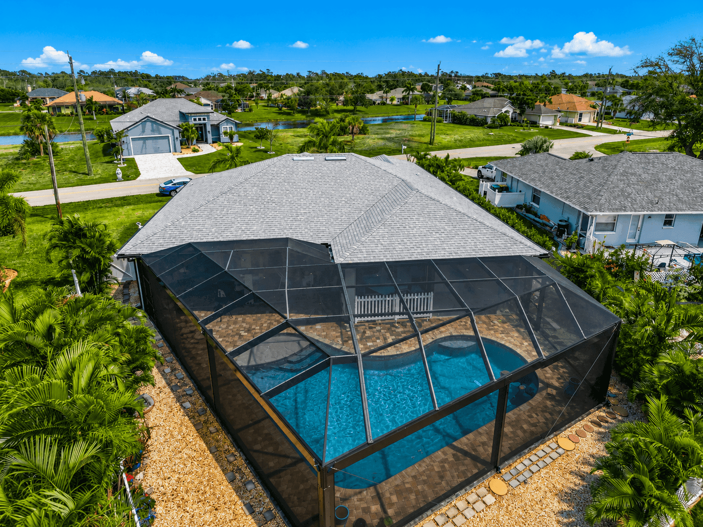 Aerial view of a house with a screened-in swimming pool in the foreground, surrounded by palm trees and single-family homes with gray roofs.